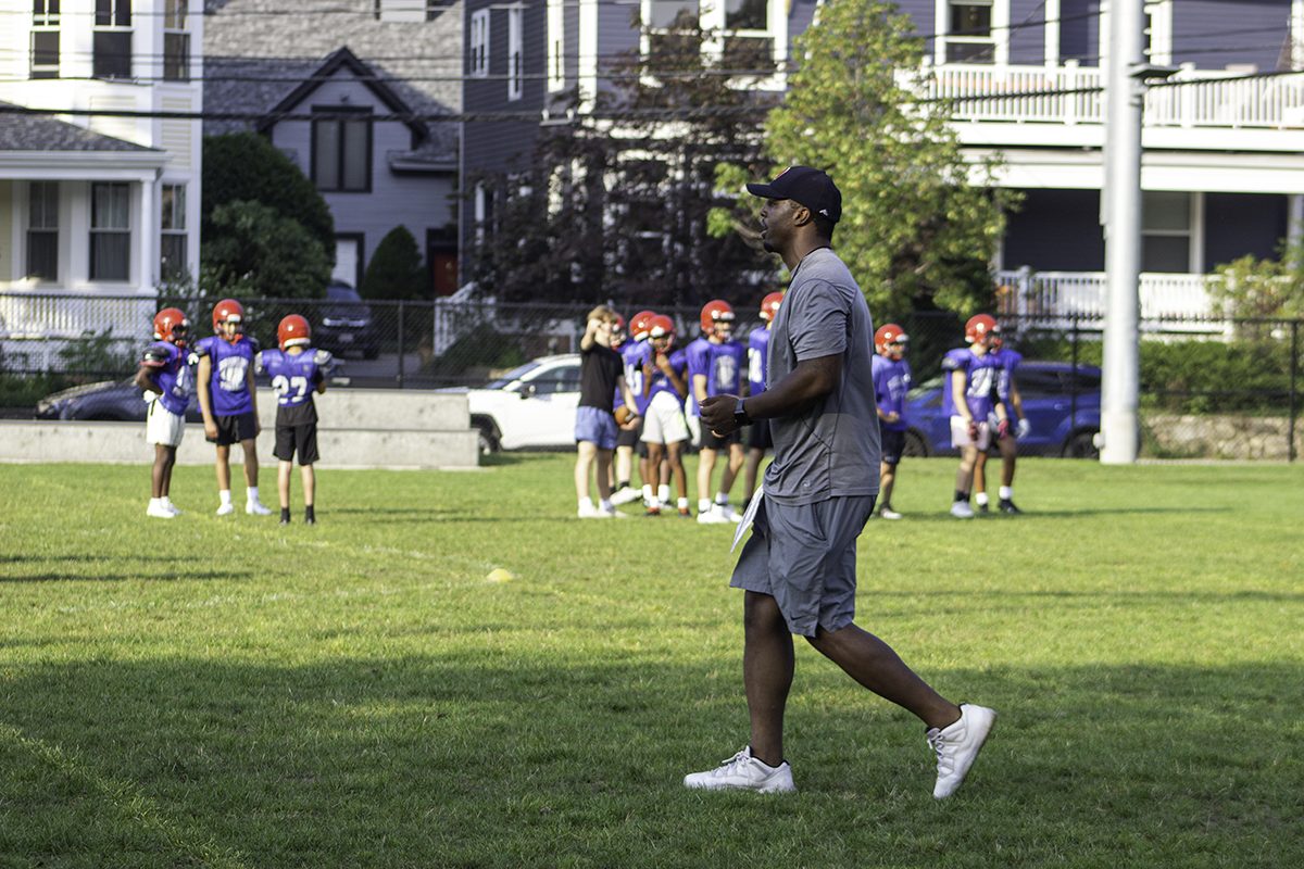 In the foreground, Brookline High School football coach Chad Hunte walks across the practice field. BHS football players stand in the background
