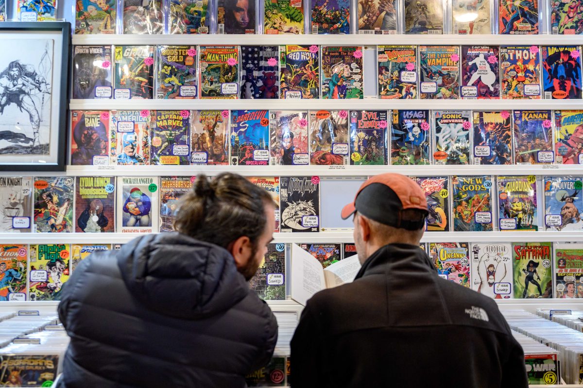 Two men seen from behind read a comic book in front of a wall covered with more comic books. 