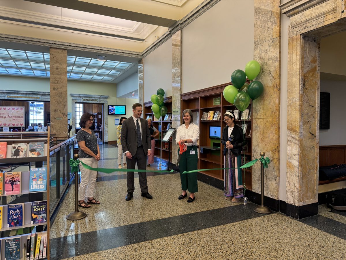 A smiling woman in green pants and a white blouse uses an oversize pair of red scissors to cut a green ribbon.