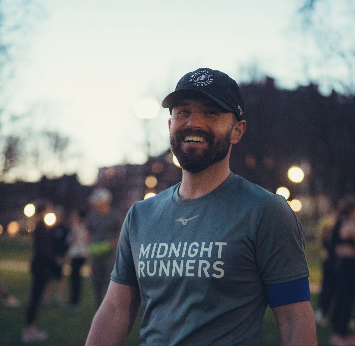 A white man with a dark beard stands, smiling, at dusk. He is wearing a baseball cap and blue t-shirt that reads "Midnight Runners."